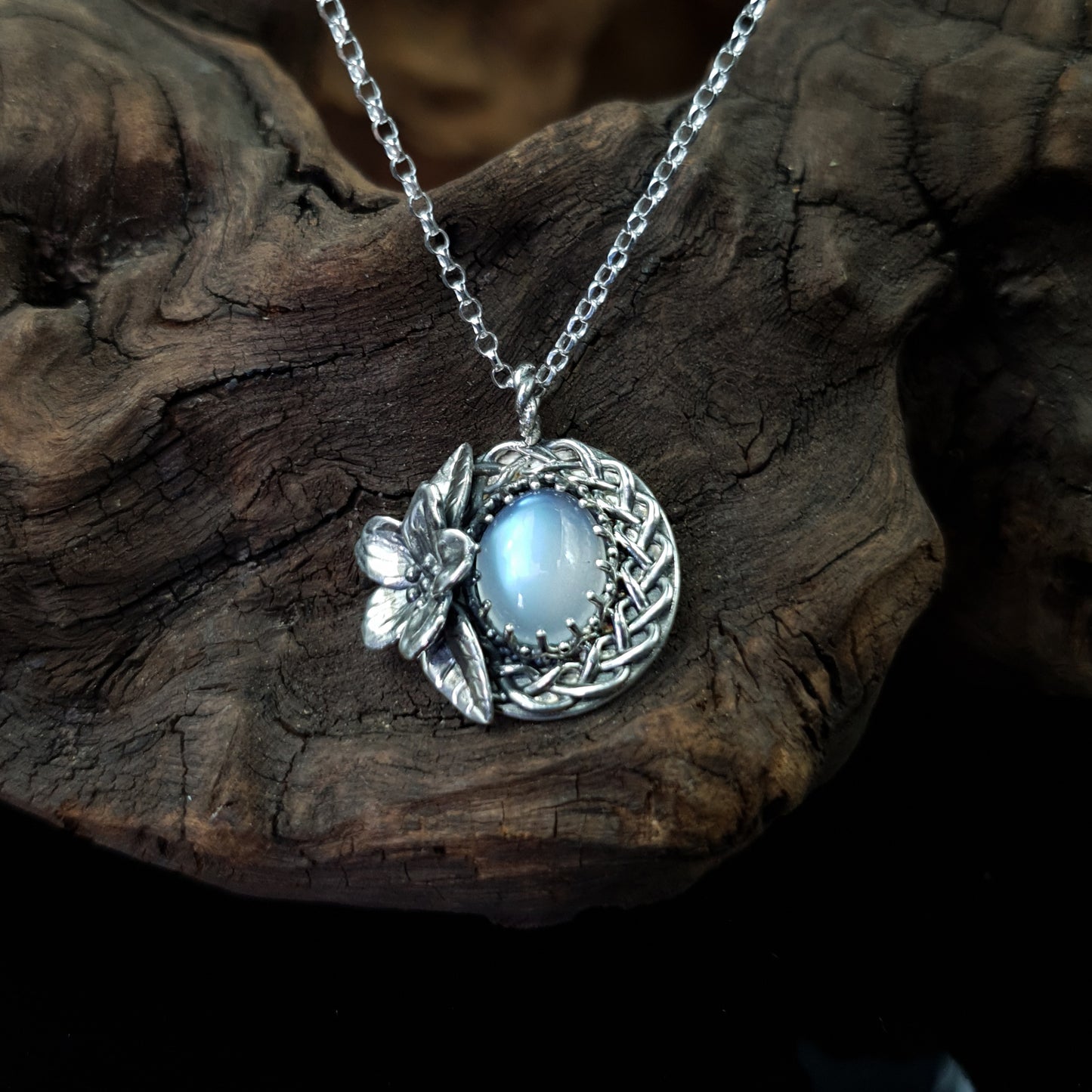 Ciondolo treccia in argento 925 con fiore di ciliegio e adularia, design elegante e artigianale, su sfondo di legno scuro.