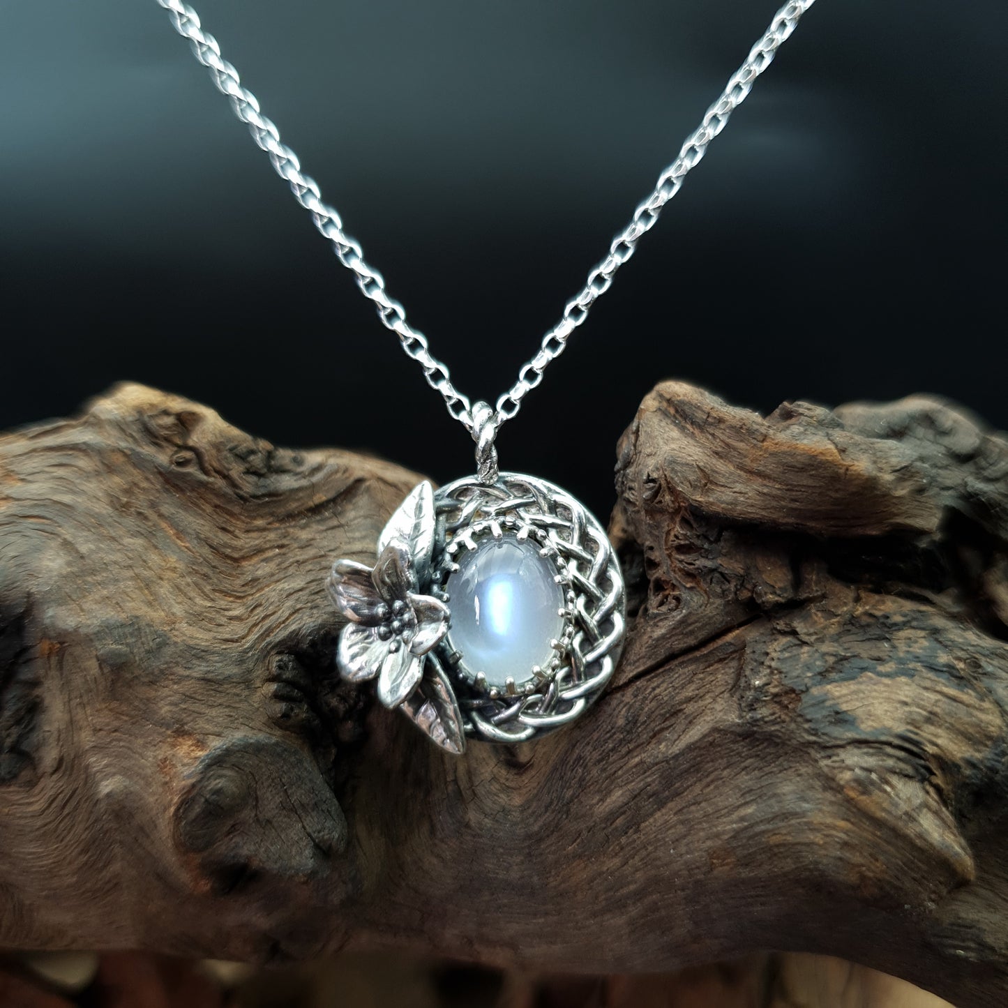 Ciondolo treccia in argento 925 con fiore di ciliegio e adularia, design elegante e artigianale, su sfondo di legno scuro.
