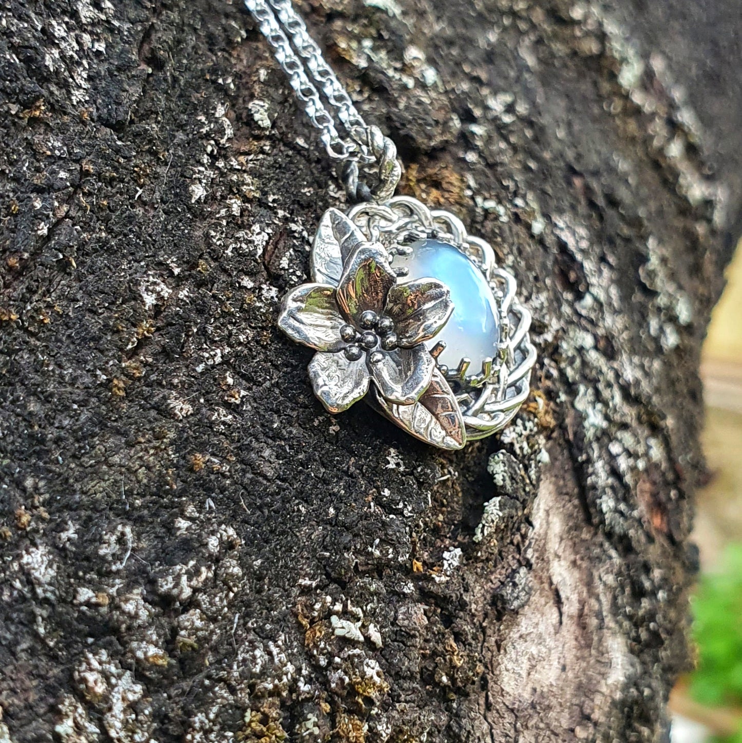 Ciondolo treccia in argento 925 con fiore di ciliegio e adularia, design elegante e artigianale, su sfondo di corteccia.