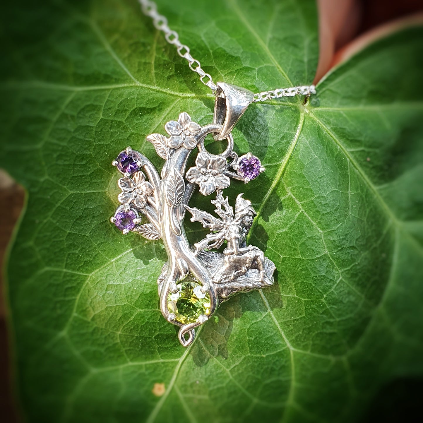 Gioiello in argento 925 con una fata seduta su una roccia, decorato con fiori e gemme di ametista viola e un grande peridoto verde.