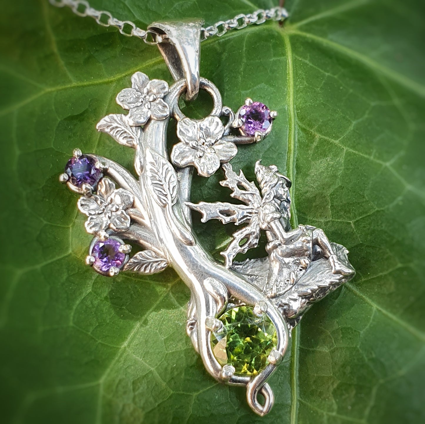 Ciondolo in argento 925 con una fata seduta su una roccia, decorato con fiori e gemme di ametista viola e un grande peridoto verde, appoggiato su una foglia verde.