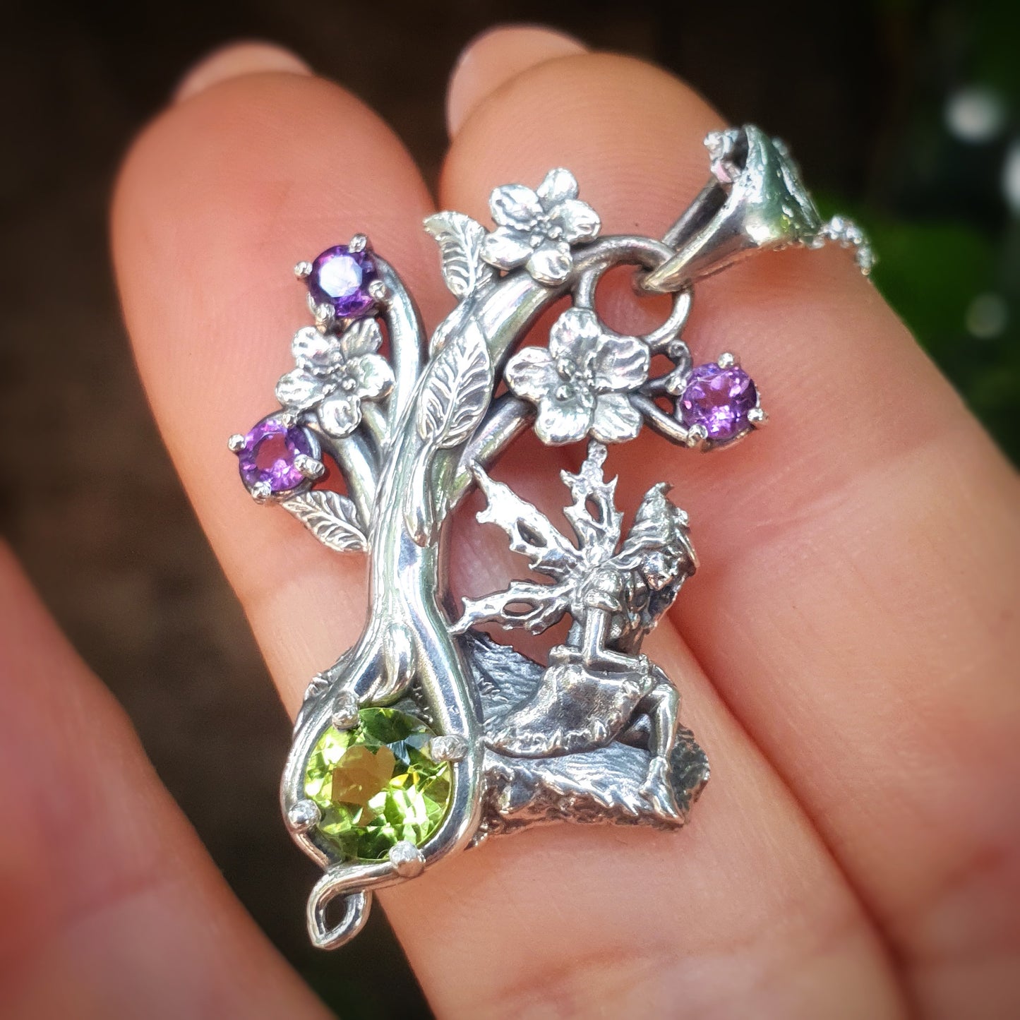 Pendente in argento 925 con una fata seduta su una roccia, decorato con fiori e gemme di ametista viola e un grande peridoto verde, tenuto tra le dita di una mano.