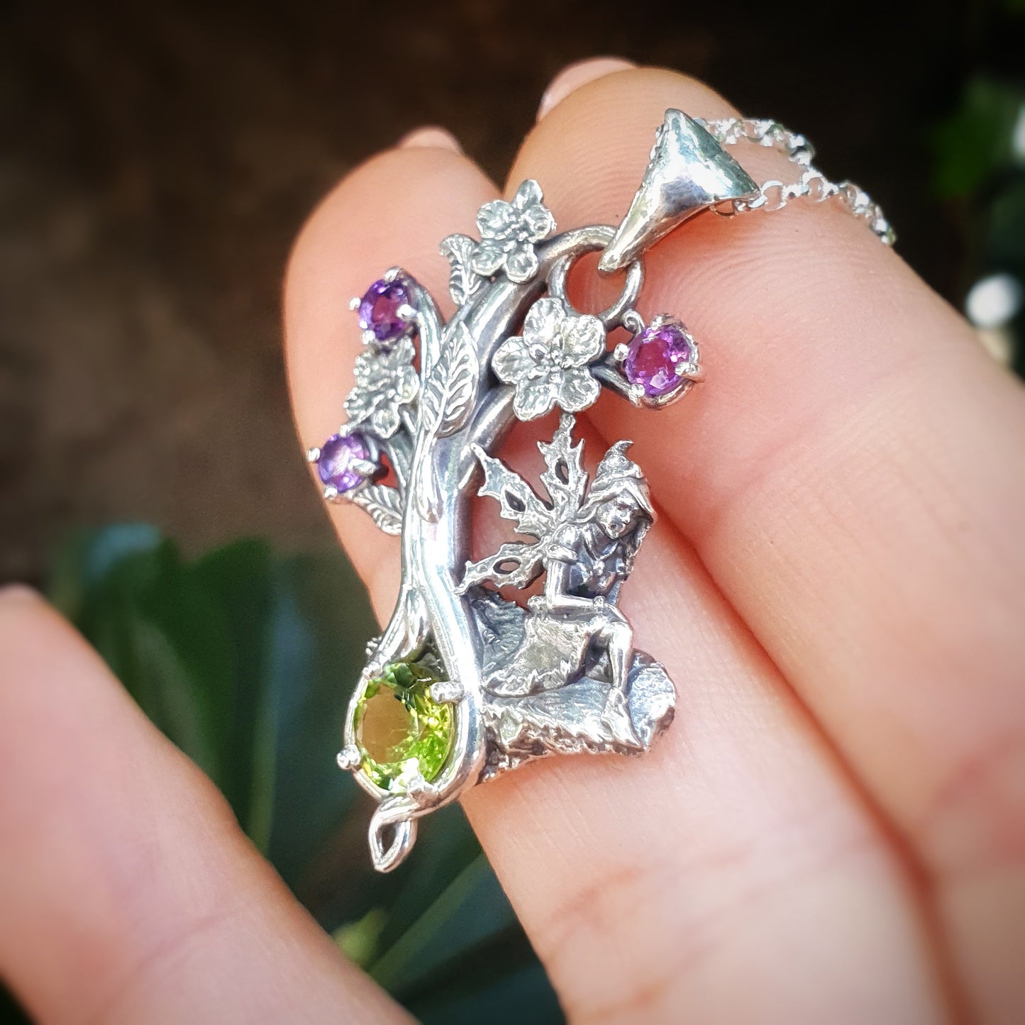Collana in argento 925 con una fata seduta su una roccia, decorata con fiori e gemme di ametista viola e un grande peridoto verde, tenuta tra le dita di una mano.