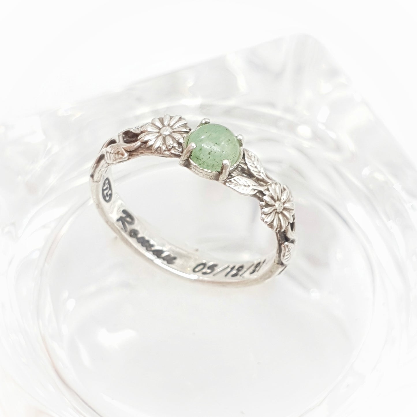 Una fede artigianale in argento poggiata su un supporto in vetro trasparente: presenta un design raffinato con un gambo lavorato, adornato da due margherite che incorniciano un cabochon centrale di avventurina verde.