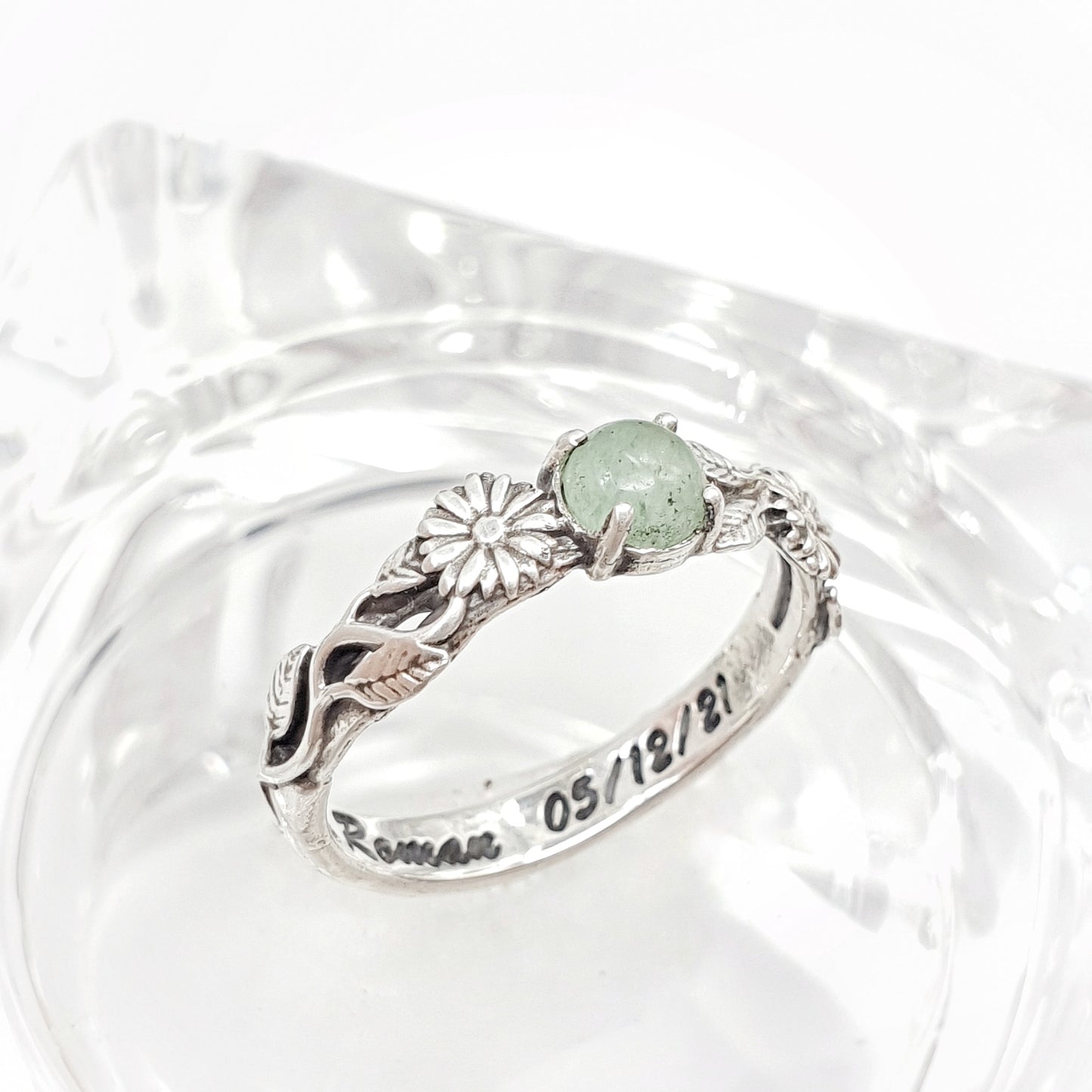 Una fede artigianale in argento poggiata su un supporto in vetro trasparente: presenta un design raffinato con un gambo lavorato, adornato da due margherite che incorniciano un cabochon centrale di avventurina verde.