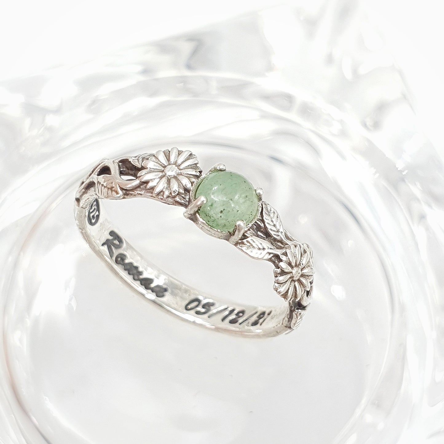 Una fede artigianale in argento poggiata su un supporto in vetro trasparente: presenta un design raffinato con un gambo lavorato, adornato da due margherite che incorniciano un cabochon centrale di avventurina verde.