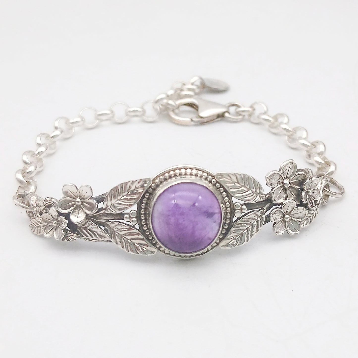 Bracciale in argento925 e ametista taglio cabochon, con piastrina rigida superiore ornata da rametti con foglie e fiori, e catenina con moschettone sul lato posteriore.