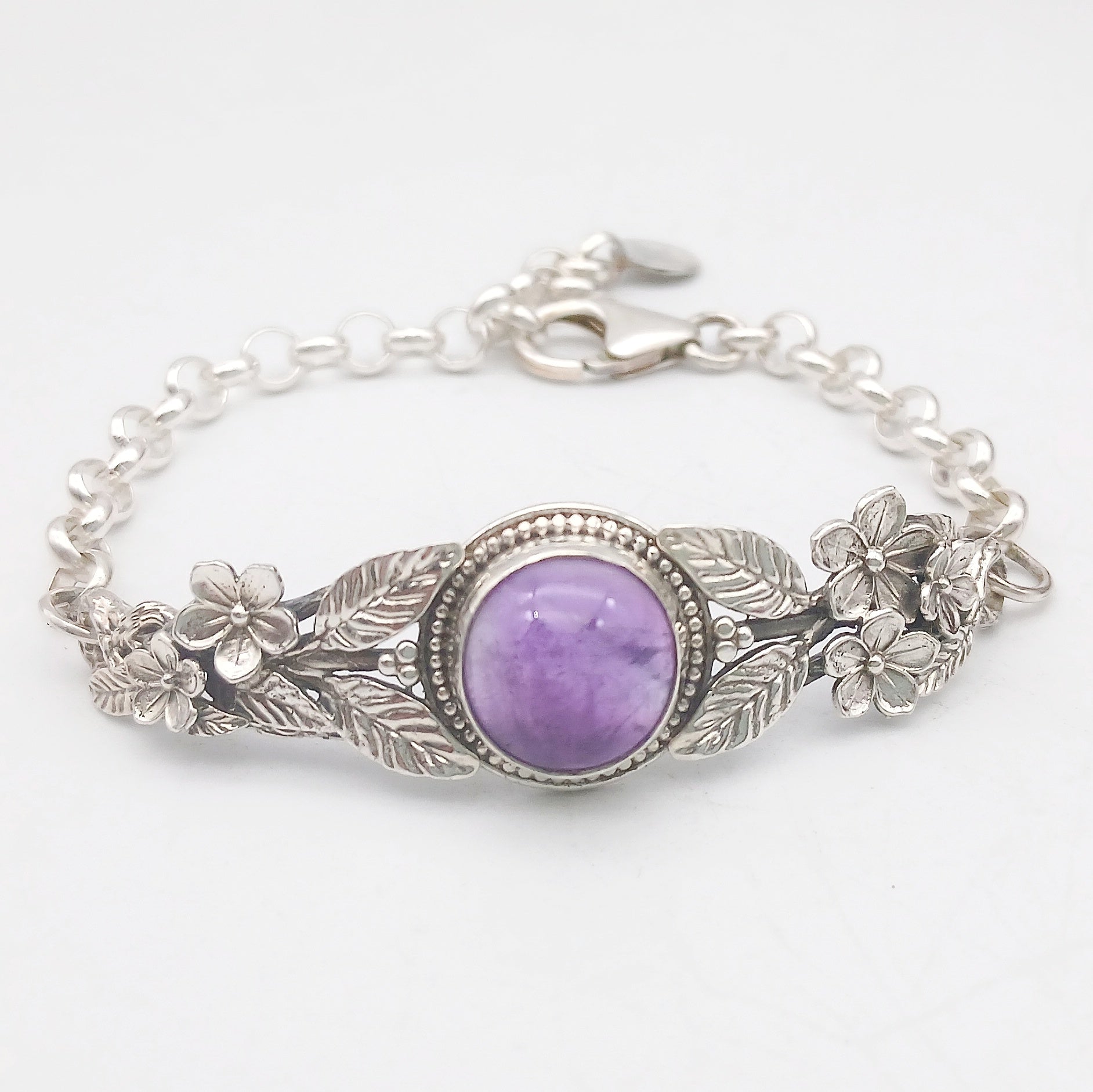 Bracciale in argento925 e ametista taglio cabochon, con piastrina rigida superiore ornata da rametti con foglie e fiori, e catenina con moschettone sul lato posteriore.
