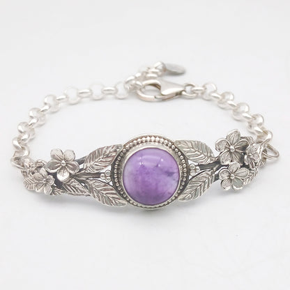 Bracciale in argento925 e ametista taglio cabochon, con piastrina rigida superiore ornata da rametti con foglie e fiori, e catenina con moschettone sul lato posteriore.