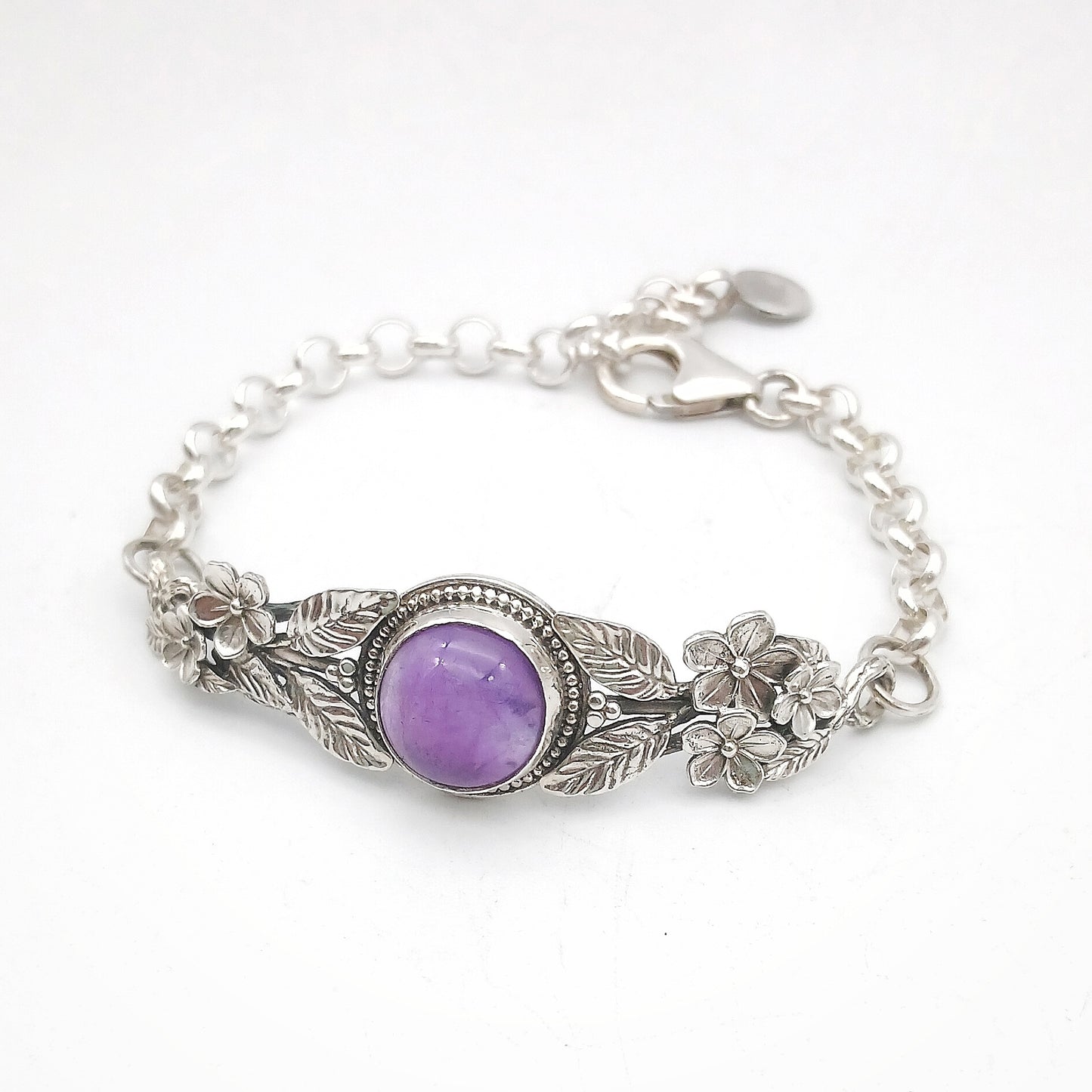 Bracciale in argento925 e ametista taglio cabochon, con piastrina rigida superiore ornata da rametti con foglie e fiori, e catenina con moschettone sul lato posteriore.