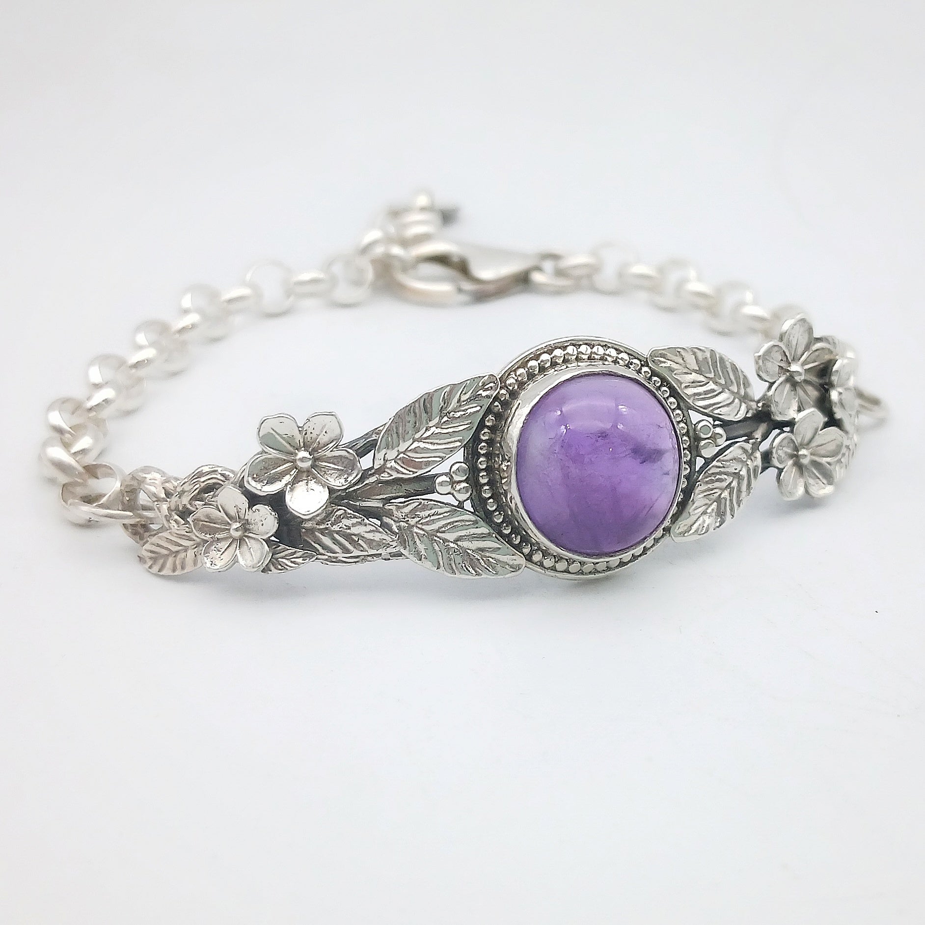 Bracciale in argento925 e ametista taglio cabochon, con piastrina rigida superiore ornata da rametti con foglie e fiori, e catenina con moschettone sul lato posteriore.