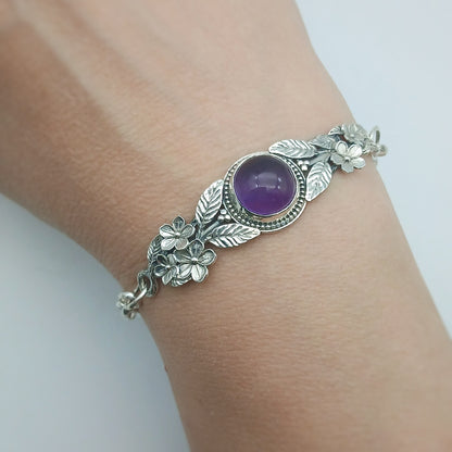 Bracciale in argento925 e ametista taglio cabochon, con piastrina rigida superiore ornata da rametti con foglie e fiori, e catenina con moschettone sul lato posteriore.
