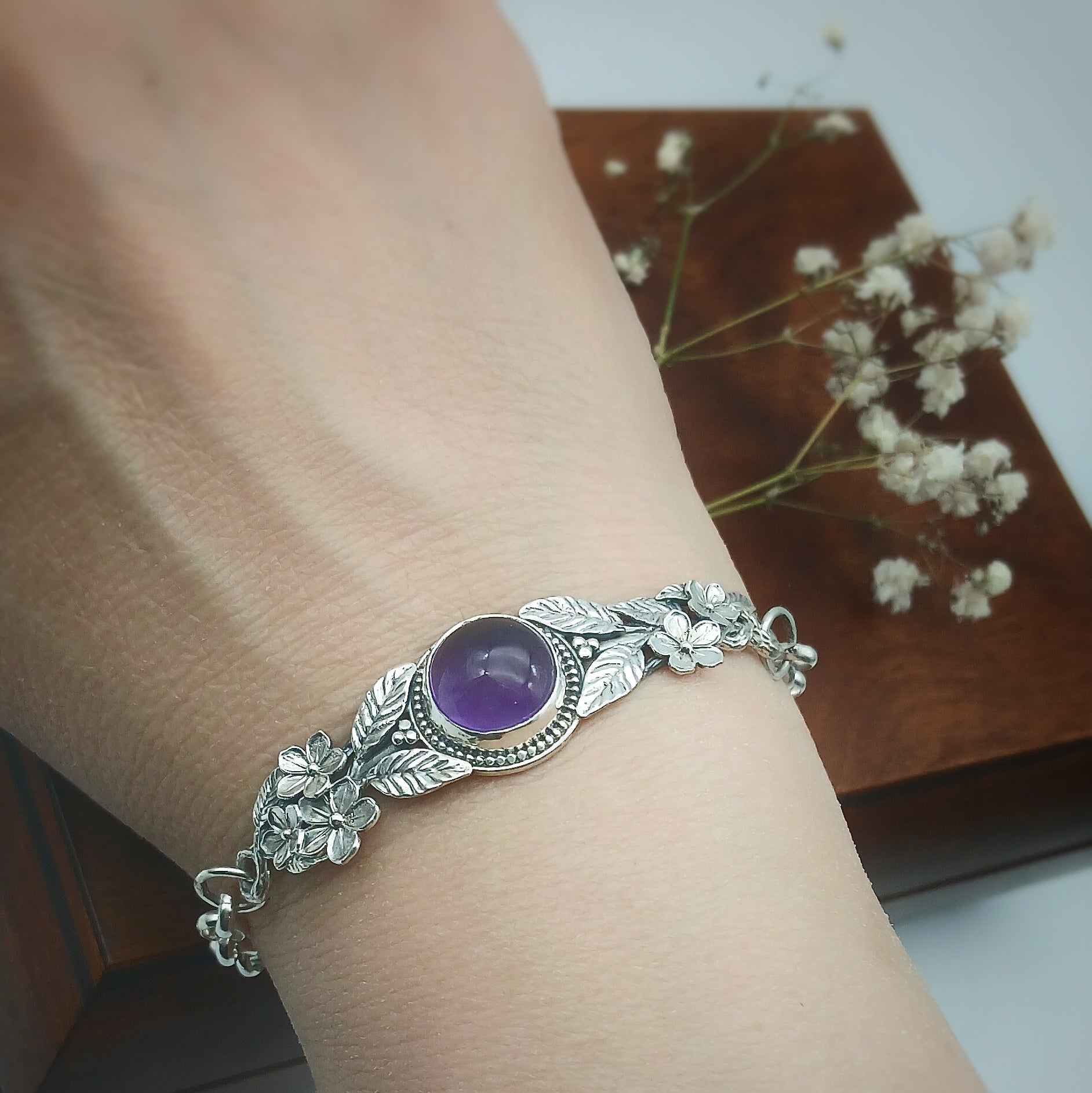 Bracciale in argento925 e ametista taglio cabochon, con piastrina rigida superiore ornata da rametti con foglie e fiori, e catenina con moschettone sul lato posteriore.