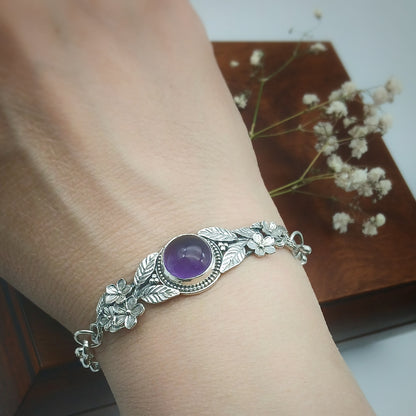 Bracciale in argento925 e ametista taglio cabochon, con piastrina rigida superiore ornata da rametti con foglie e fiori, e catenina con moschettone sul lato posteriore.