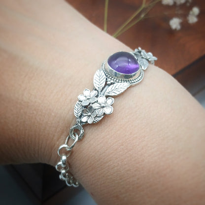 Bracciale in argento925 e ametista taglio cabochon, con piastrina rigida superiore ornata da rametti con foglie e fiori, e catenina con moschettone sul lato posteriore.