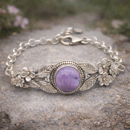 Bracciale in argento925 e ametista taglio cabochon, con piastrina rigida superiore ornata da rametti con foglie e fiori, e catenina con moschettone sul lato posteriore.