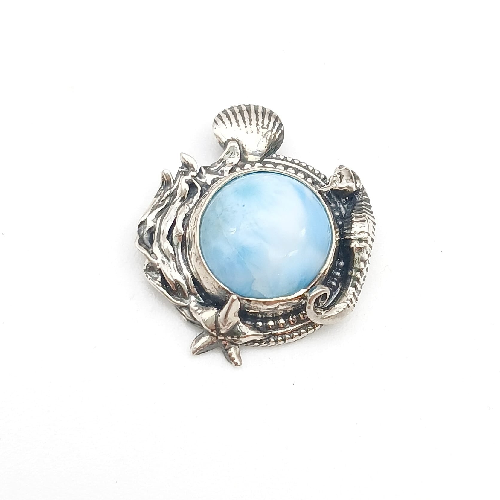 Ciondolo in argento 925 e cabochon di larimar rotondo, circondato da decorazioni di fondale marino, alghe, conchiglie, stella marina e un cavalluccio marino sul lato destro.