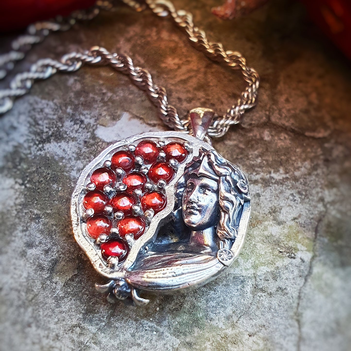 Ciondolo in argento 925 raffigurante una melagrana divisa a metà: a sinistra dodici granati rossi da 3mm, a destra una microscultura della dea Persefone.