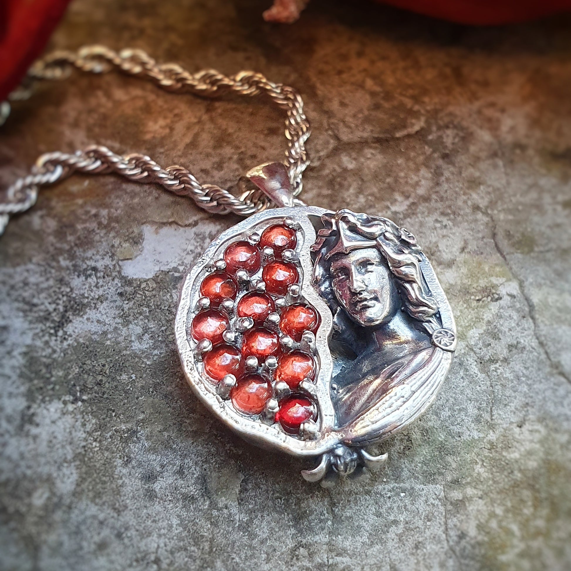Ciondolo in argento 925 raffigurante una melagrana divisa a metà: a sinistra dodici granati rossi da 3mm, a destra una microscultura della dea Persefone.