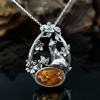 Ciondolo in argento raffigurante una volpe seduta su un cabochon ovale di ambra baltica. La volpe è sormontata da due rami che la avvolgono, i rami sono adorni di fiori e foglie.