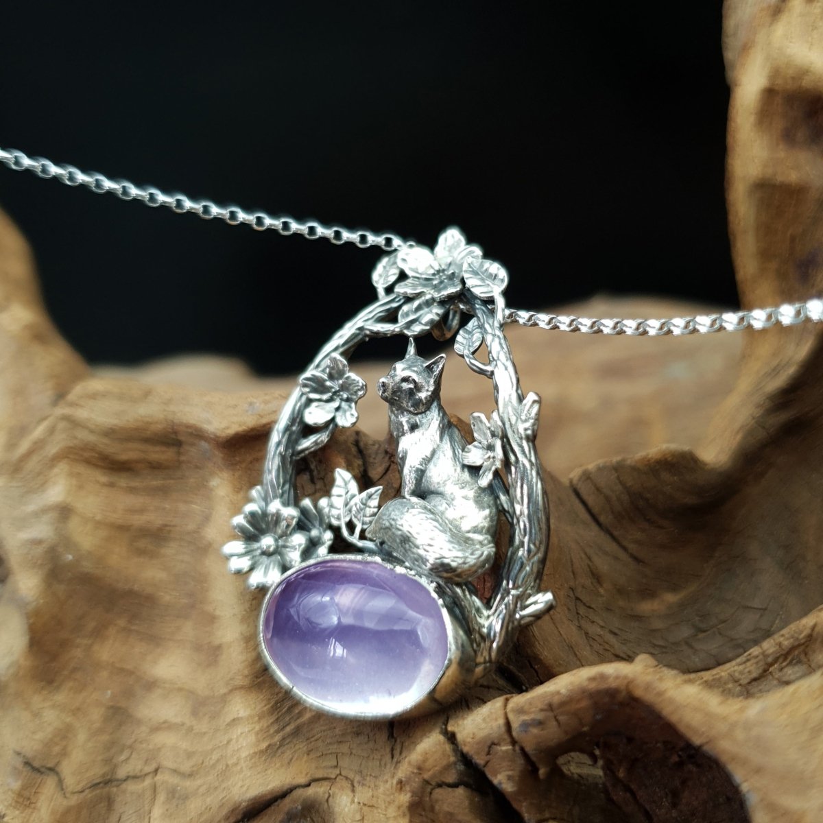Ciondolo in argento raffigurante una volpe seduta su un cabochon ovale di ametista. La volpe è sormontata da due rami che la avvolgono, i rami sono adorni di fiori e foglie.
