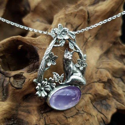 Ciondolo in argento raffigurante una volpe seduta su un cabochon ovale di ametista. La volpe è sormontata da due rami che la avvolgono, i rami sono adorni di fiori e foglie.