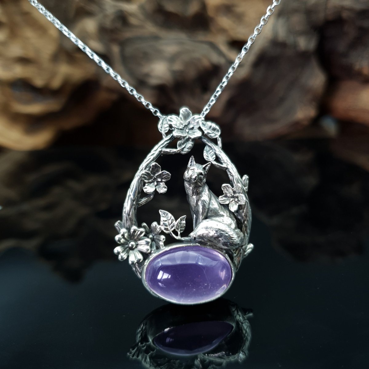 Ciondolo in argento raffigurante una volpe seduta su un cabochon ovale di ametista. La volpe è sormontata da due rami che la avvolgono, i rami sono adorni di fiori e foglie.